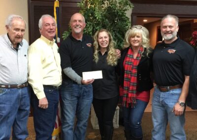 Six adults stand indoors, smiling and posing for a group photo. One woman in the center holds a white envelope—likely a donation to end childhood hunger from a Stillwater OK nonprofit charity. An American flag and potted plant are visible in the background. -Kickin' Childhood Hunger