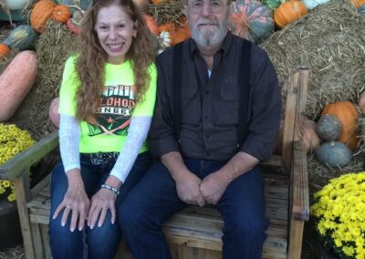 A smiling woman and man sit on a wooden bench in front of a festive fall display with pumpkins, gourds, hay bales, and yellow flowers—capturing the spirit of giving in Stillwater OK charity events supporting donations to help hungry children. -Kickin' Childhood Hunger