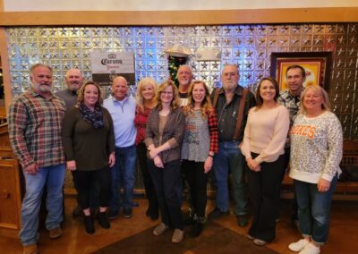 A group of thirteen adults stand close together, smiling at the camera inside a restaurant with a glass block wall and framed artwork—gathered to support a Stillwater OK charity working to end childhood hunger in their community. -Kickin' Childhood Hunger