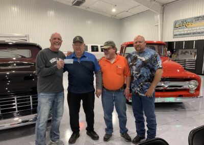 Four men stand smiling in a garage between two classic trucks; one man holds a check. The shiny floors and high ceiling frame a joyful moment as their Stillwater OK charity works to help end childhood hunger. -Kickin' Childhood Hunger