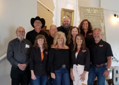 A group of ten adults, smiling and posing indoors. Most wear black tops with a Stillwater OK charity logo, and one man sports a black cowboy hat. They stand before a white draped background and a decorative sign. -Kickin' Childhood Hunger