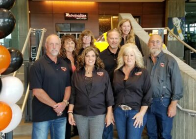 A group of eight adults, casually dressed, stand together and smile for a photo indoors near a staircase. With orange, black, and white balloons nearby and a Metropolitan Imported sign behind them, this Stillwater OK charity works to end childhood hunger. -Kickin' Childhood Hunger