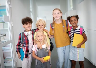Five smiling children with backpacks stand together in a hallway, holding notebooks and folders, looking happy and ready for school—thanks to a Stillwater OK charity that helps hungry children and works to end childhood hunger in the community. -Kickin' Childhood Hunger