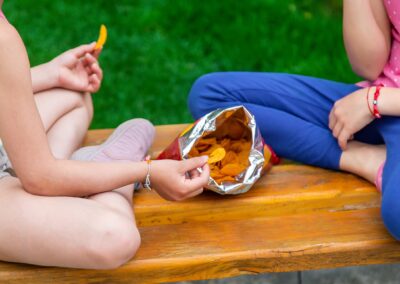 Two children sit cross-legged on a wooden bench, sharing a bag of chips. Both reach into the open bag, green grass behind them—a simple moment that highlights why Stillwater OK nonprofit charity efforts work to end childhood hunger. -Kickin' Childhood Hunger