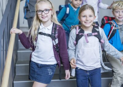 A group of young children with backpacks walk down a staircase together, smiling and chatting. Two girls lead the group, holding the handrail—just one scene where a local charity helps hungry children in their Stillwater OK school hallway. -Kickin' Childhood Hunger