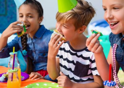 Three children sit at a colorful table, wearing party hats and eating decorated cupcakes, smiling and enjoying a festive celebration—made possible by a local charity that helps hungry children in Stillwater, OK. -Kickin' Childhood Hunger