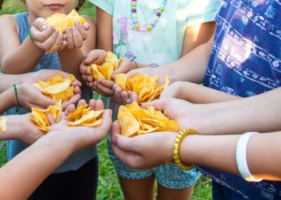 A group of children standing outdoors in a circle, each holding a handful of potato chips and sharing them with one another—an uplifting moment made possible as donations help hungry children through a Stillwater OK charity. -Kickin' Childhood Hunger