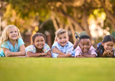 Five young children of diverse backgrounds lie on green grass, smiling and laughing together on a sunny day. Donations help hungry children like these find joy—support a Stillwater OK charity to make a difference today. -Kickin' Childhood Hunger