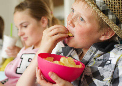 A boy wearing a straw hat eats potato chips from a pink bowl, while a girl in the background drinks from a cup with a straw. They appear to be indoors, with bookshelves behind them—reminding us how local charity helps hungry children. -Kickin' Childhood Hunger