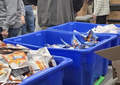 People are gathered around tables filled with blue bins containing snack bags. Cardboard boxes are stacked nearby, and several people are working together in a room, possibly packing food or organizing supplies. -Kickin' Childhood Hunger