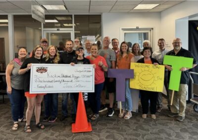 A group of smiling people pose indoors holding a large check, colorful signs including a sun and “Welcome Home Sunshine!” The check is made out to “Kickin Childhood Hunger” for $120,000. -Kickin' Childhood Hunger