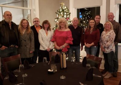 A group of ten adults stand together and smile for a photo in a restaurant with a round table in the foreground and a decorated Christmas tree in the background. -Kickin' Childhood Hunger