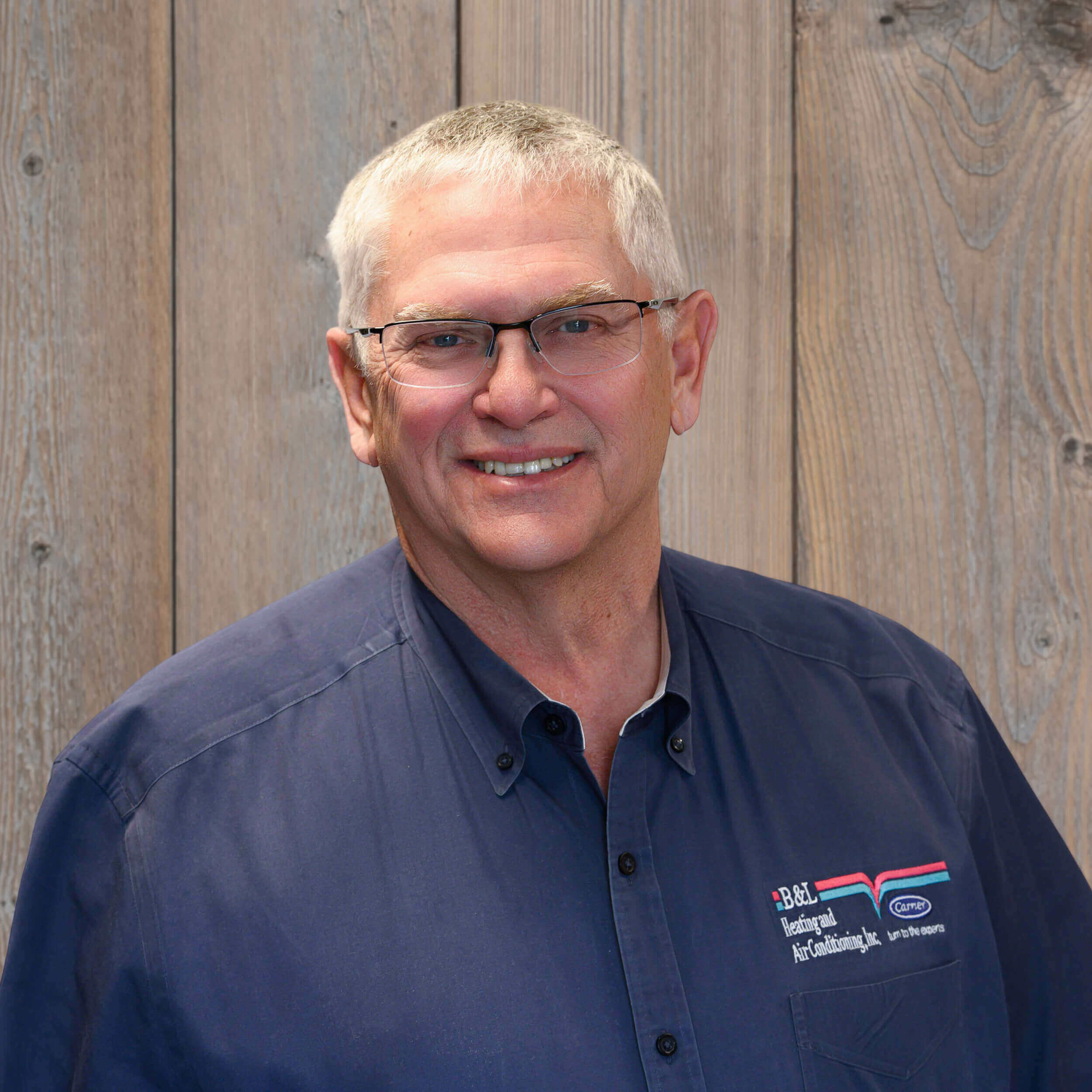 Smiling older man with short gray hair and glasses wearing a navy blue button-up shirt with a company logo, standing in front of a wooden plank background. -Kickin' Childhood Hunger